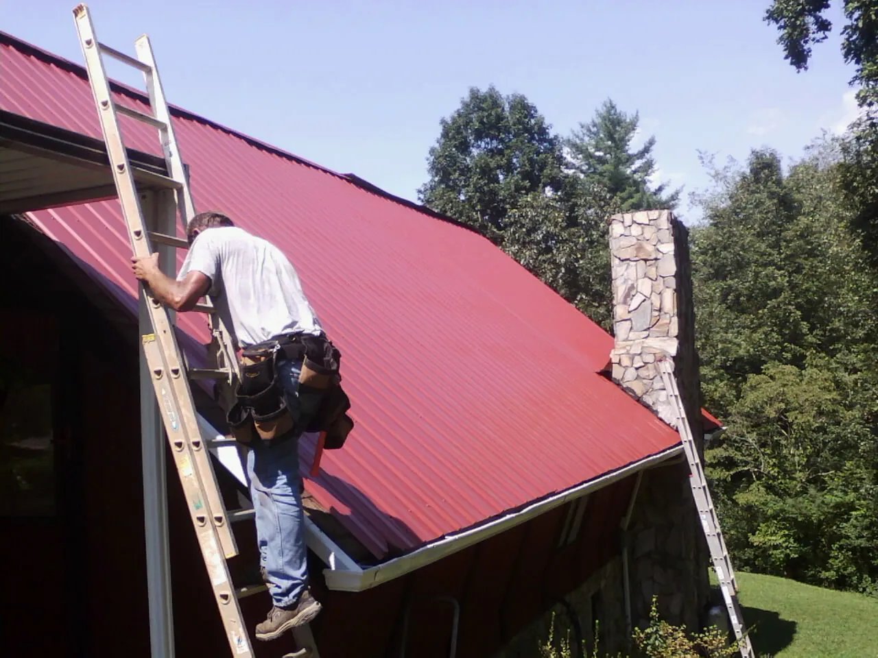 Roofer on a ladder beside a red metal roof and stone chimney — Soffit & Fascia Repair by Roofing Brothers in Turpin
